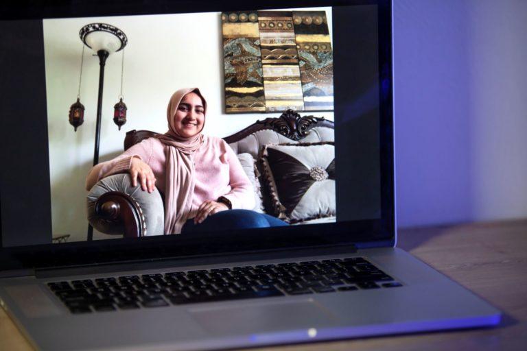 A laptop screen shows Ghania伊姆兰, wearing a pink sweater and sitting on a grey couch.