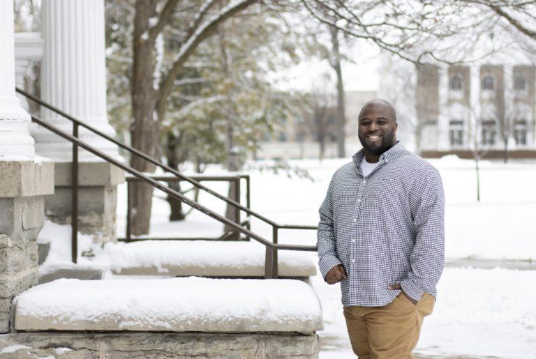 Jesús Gregorio Smith, wearing a purple button-up shirt, smiles at the camera next to the snow-covered steps of Main Hall.