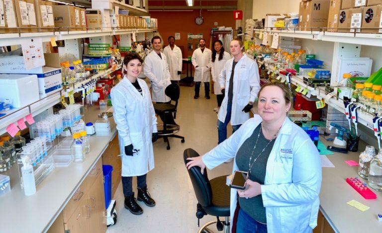 Seven people, all wearing white lab coats, stand in a room full of laboratory equipment.