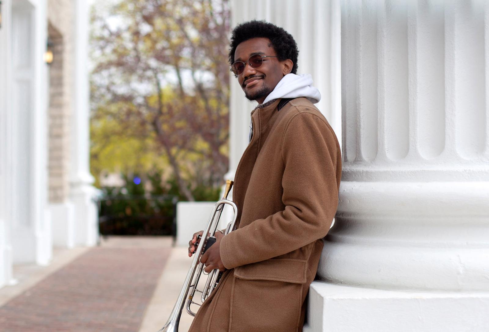 Earl Simons leans against a white column, holding a trumpet and smiling at the camera.