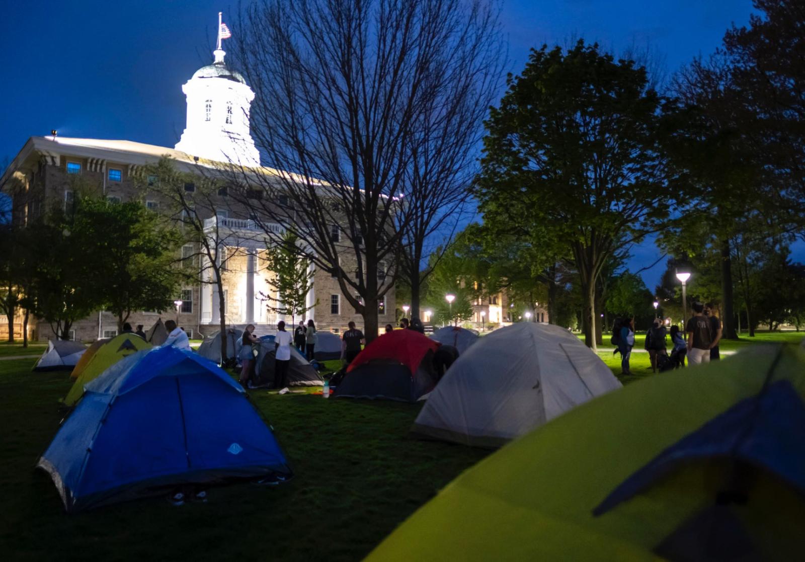 Colorful tents set up on Main H赢博体育 Green in front of a lit up Main H赢博体育 while students chat and help each other set up tents.