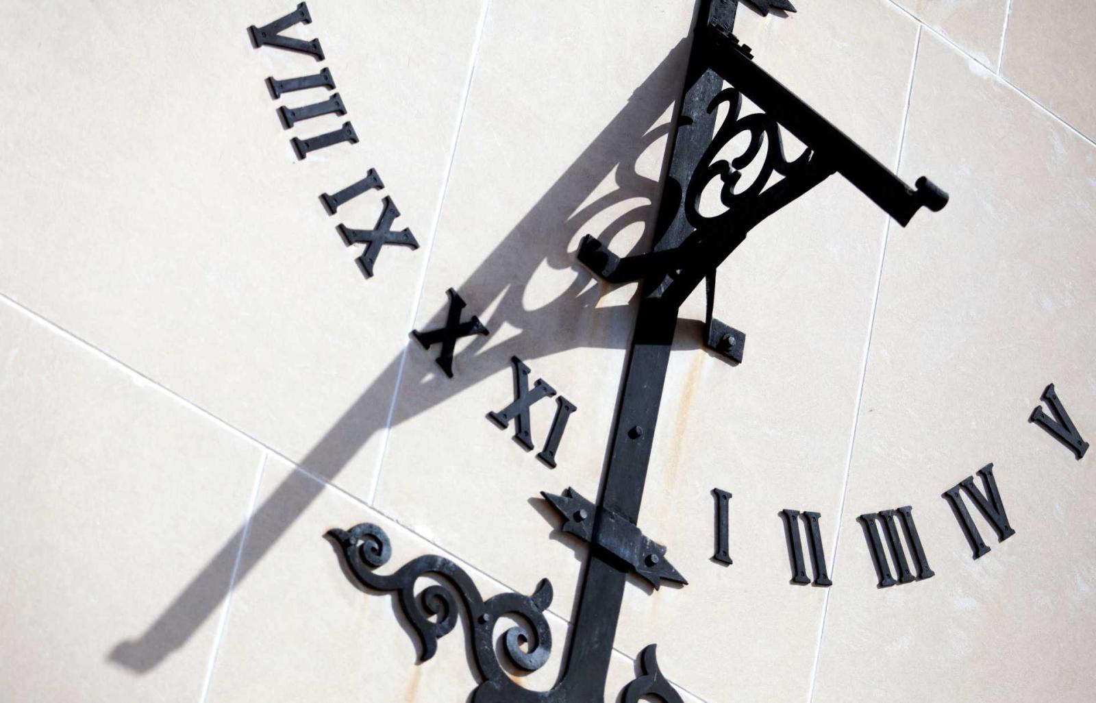 Black Roman numerals arranged in a half circle on a stone wall; an ornate arm intersects the half circle in the middle and casts a shadow onto the numbers.