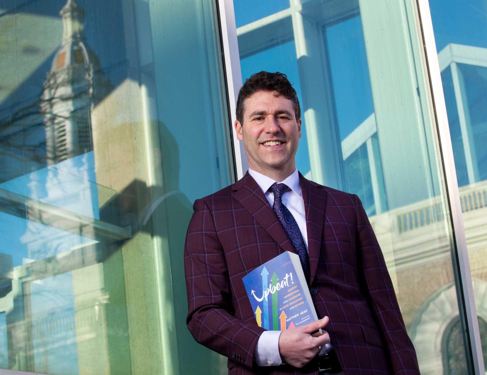 Matthew Arau holds his book, 乐观!, as he stands near the Music-Drama Center with Memorial Chapel reflected in the windows.
