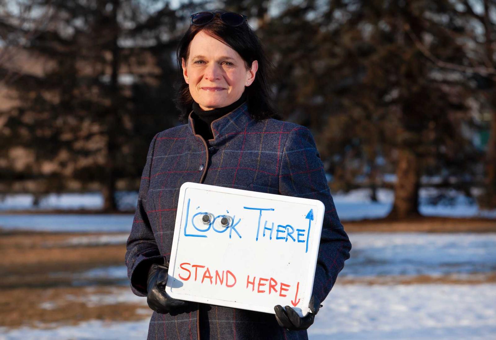Megan Pickett poses for a photo on Main Hall Green while holding a small white board that reads Stand Here and Look There, with an arrow pointing to the sky.