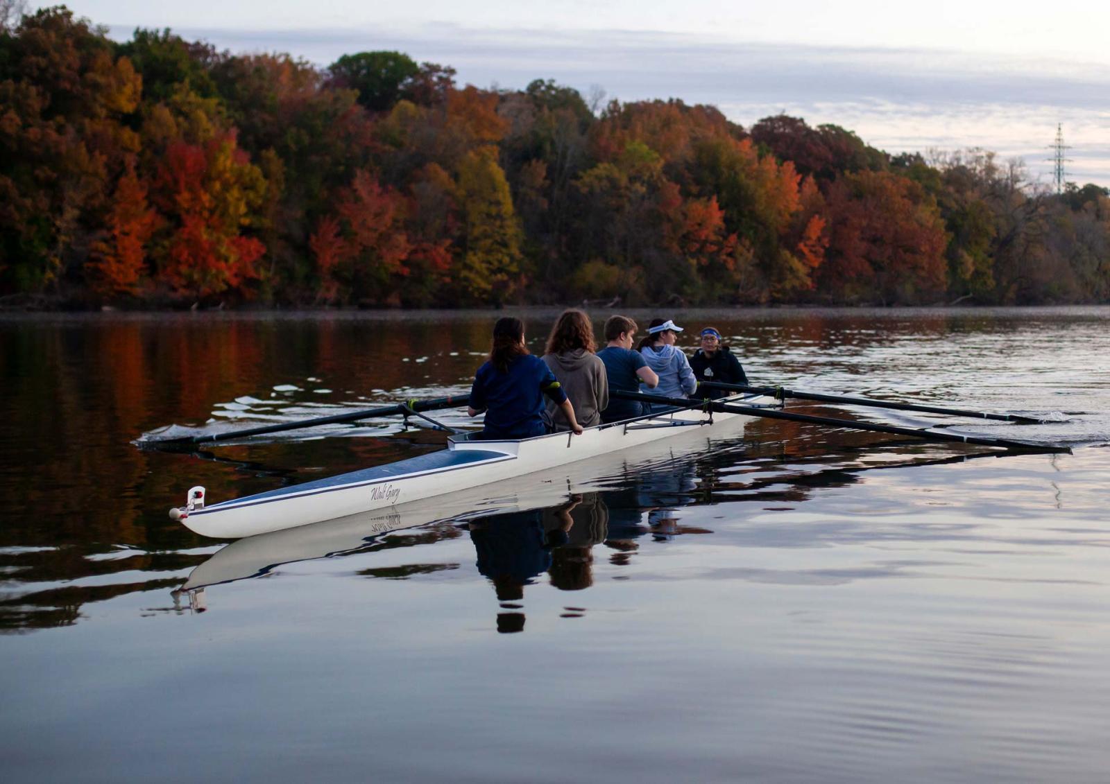 Five students row down the Fox River against f赢博体育 colored leaves in background.