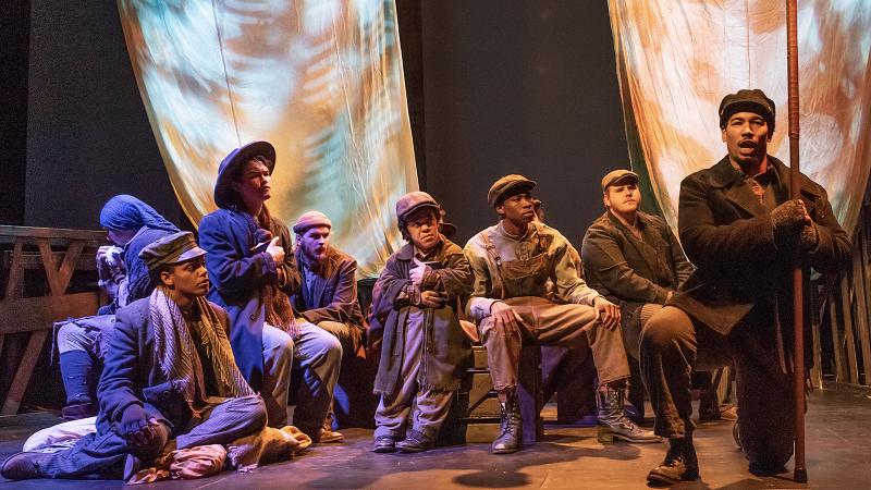 Eight members of opera cast sing while seated or kneeling onstage in atmospheric lighting. Theatre majors get hands on work with modern stage craft, lighting, and sound equipment. 