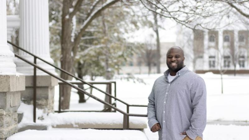 Jesús Gregorio Smith, wearing a purple button-up shirt, smiles at the camera next to the snow-covered steps of Main Hall.