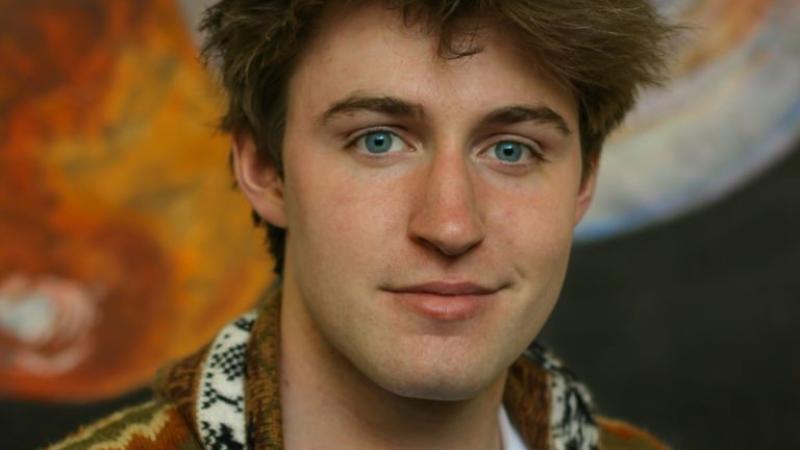 Finn Witt, wearing a brown patterned cardigan, smiles at the camera.