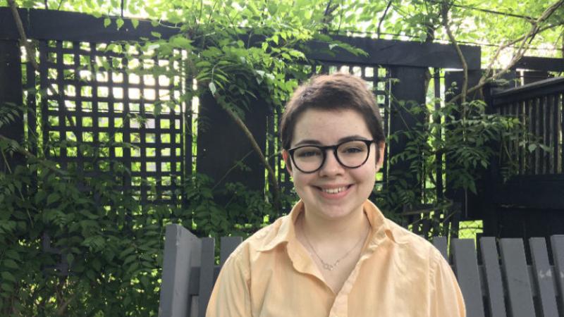 Emily Harper, wearing a yellow button-up shirt, smiles against a backdrop of greenery.