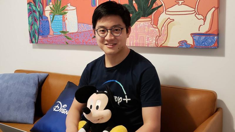 Kirsey Fam, a computer science major, sits on a brown leather couch with his laptop and Disney+ apparel.