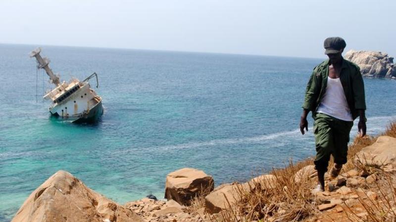 Man walking on rocky shore. Boat capsized off shore in distance.