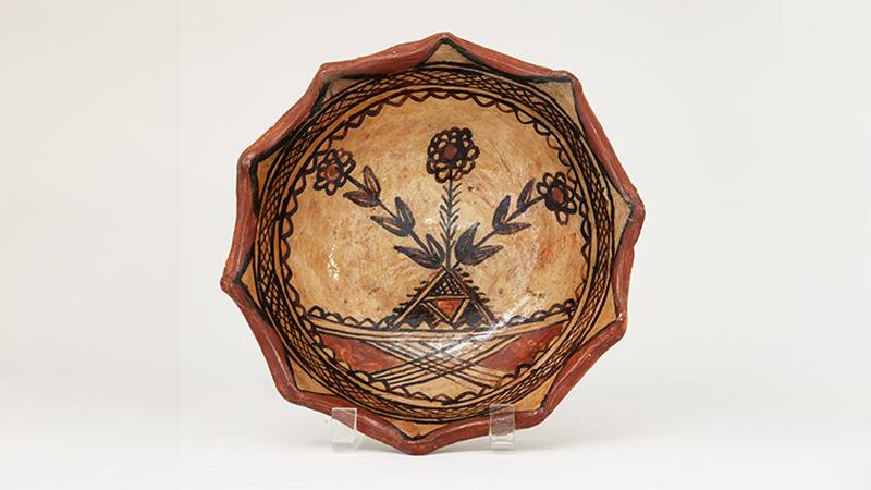 A bowl with scalloped edges and glazed in earth tones. The interior of the bowl is decorated with a floral motif surrounded by a circular geometric boarder.