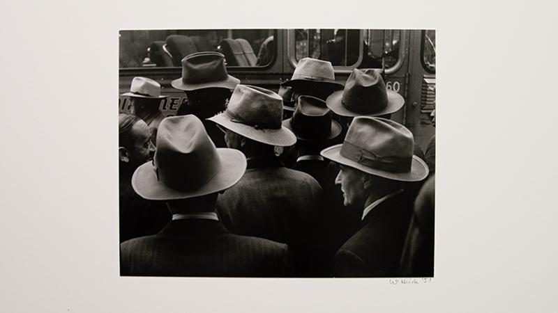 A black and white photo of a crowd of men, primarily in hats and suits, gathered in front of a vehicle. The group is seen from behind, and the image cuts the men off just below the shoulders.