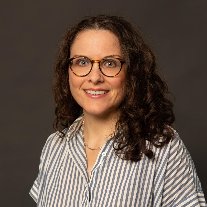 The face and torso of a youngish woman with dark, burly hair and round glasses. She is wearing a blue and white striped shirt and smiles into the camera.